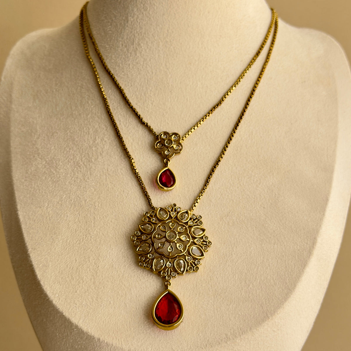 Layered gold-tone necklace with ornate pendant and red teardrop accents on display bust